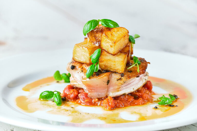 A tender slice of pork is served with roasted potatoes, a rich sauce and basil leaves.